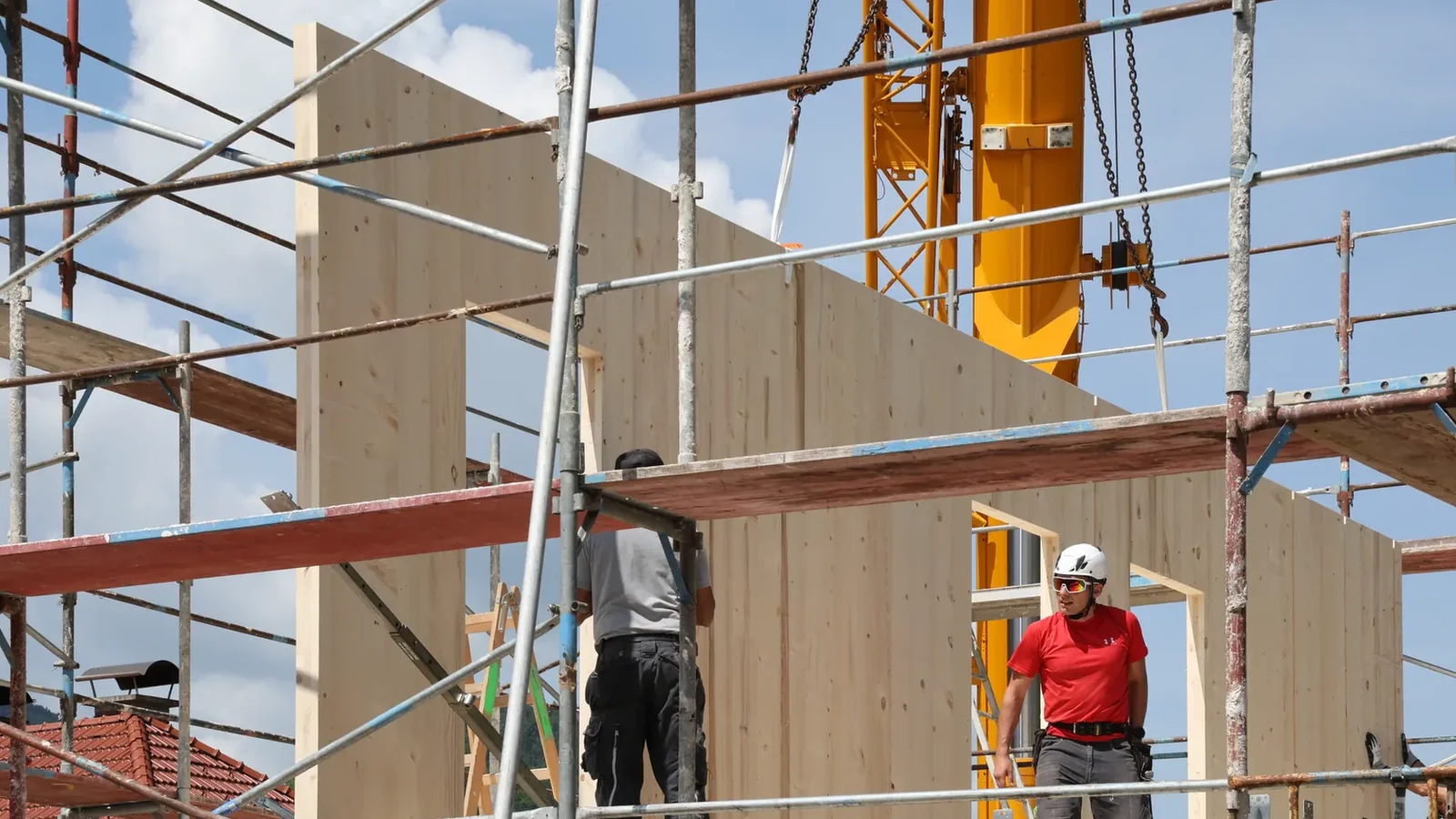 Der Kran hebt eine Wand aus Massivholz an ihren Platz, das Element hängt an den Ketten, zwei Arbeiter führen es; die erste Wand mit Fensteröffnung steht bereits