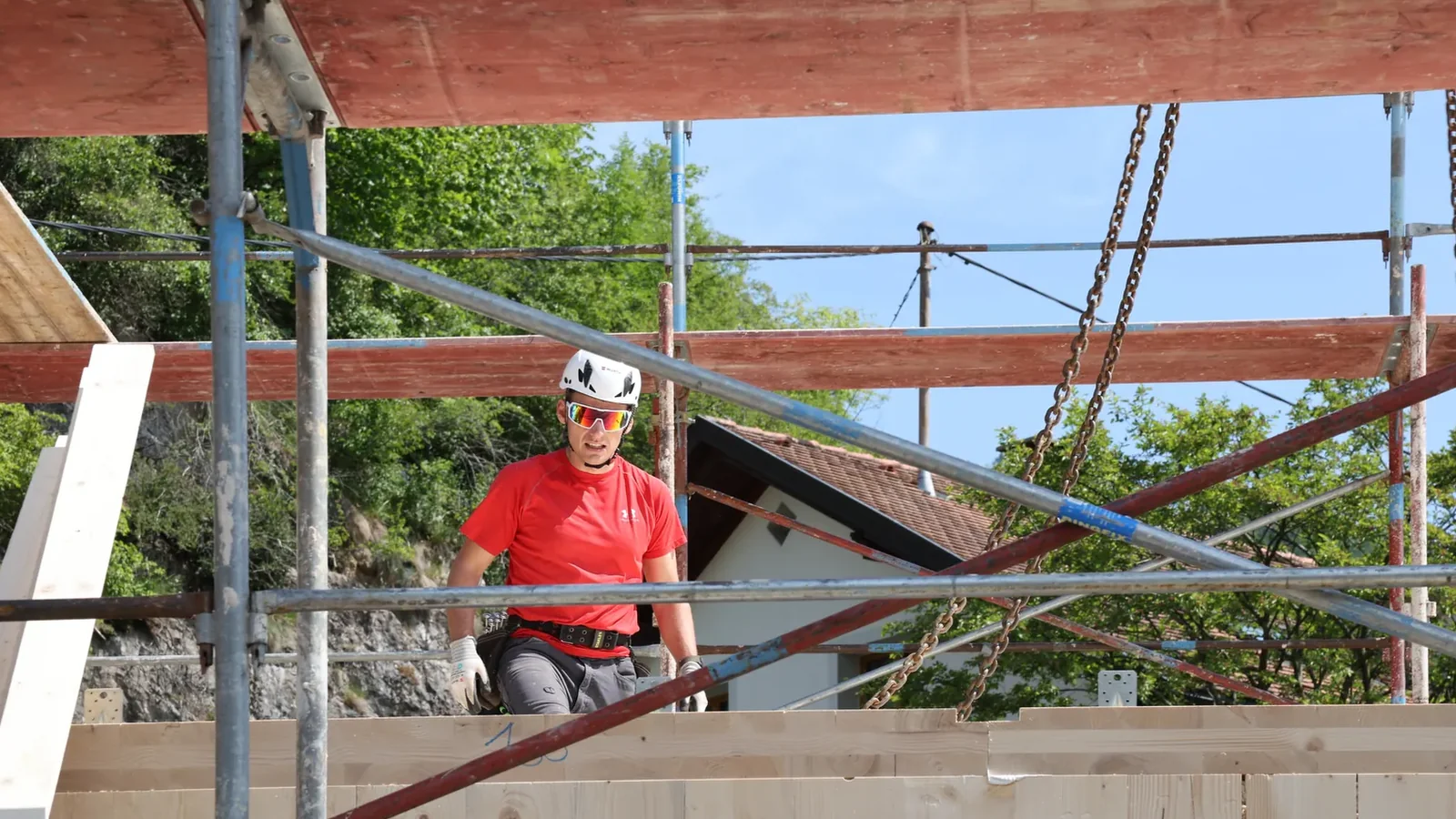Der Baumeister steht mit Helm auf der eben versetzten Massivholzwand, daneben hängen die Ketten des Krans