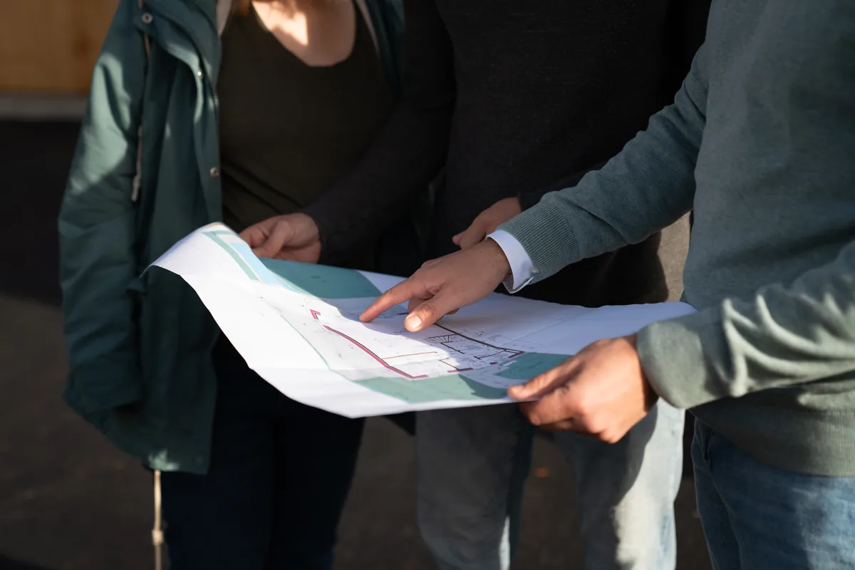 Zwei Bauherren und der Baumeister gehen gemeinsam den Plan durch, ein Finger zeigt auf das Grundstück